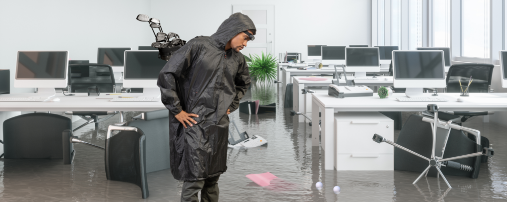 Man in a rain slicker standing in a flooded office. He has golf clubs on his back and is looking down at floating golf balls.