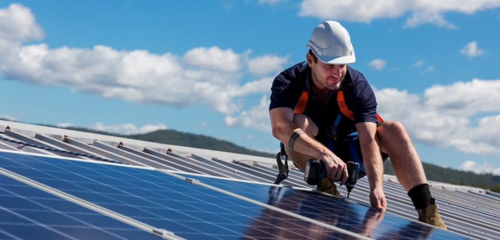 Man in white hard hat installing solar panels on a commercial building’s roof.