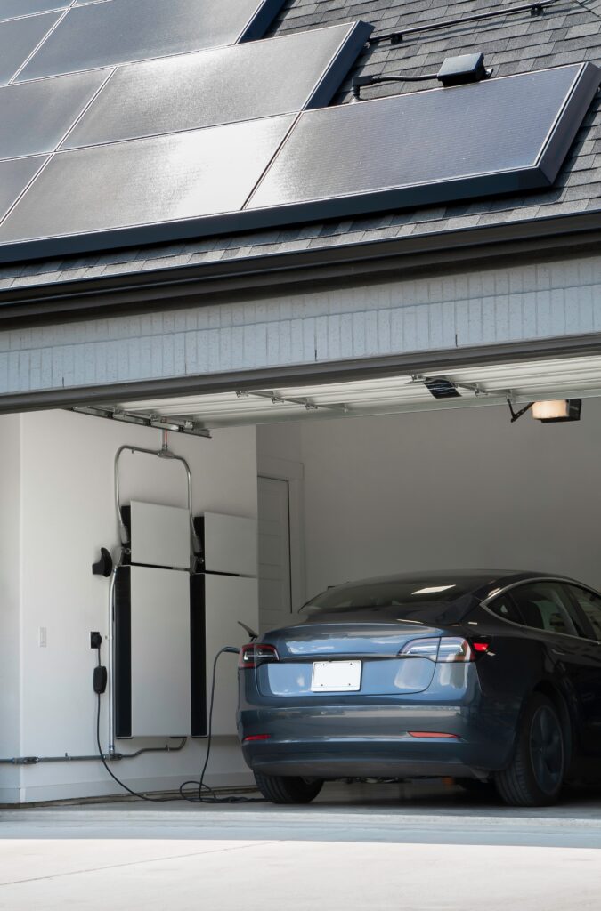 An electric vehicle sits in a garage attached to a battery backup system under the roof of a home with installed solar panels. 